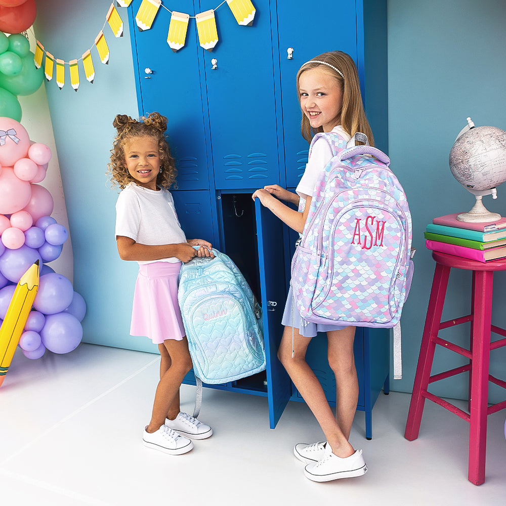 2 gilrs holding blue quilted hearts backpack and mermaid backpack