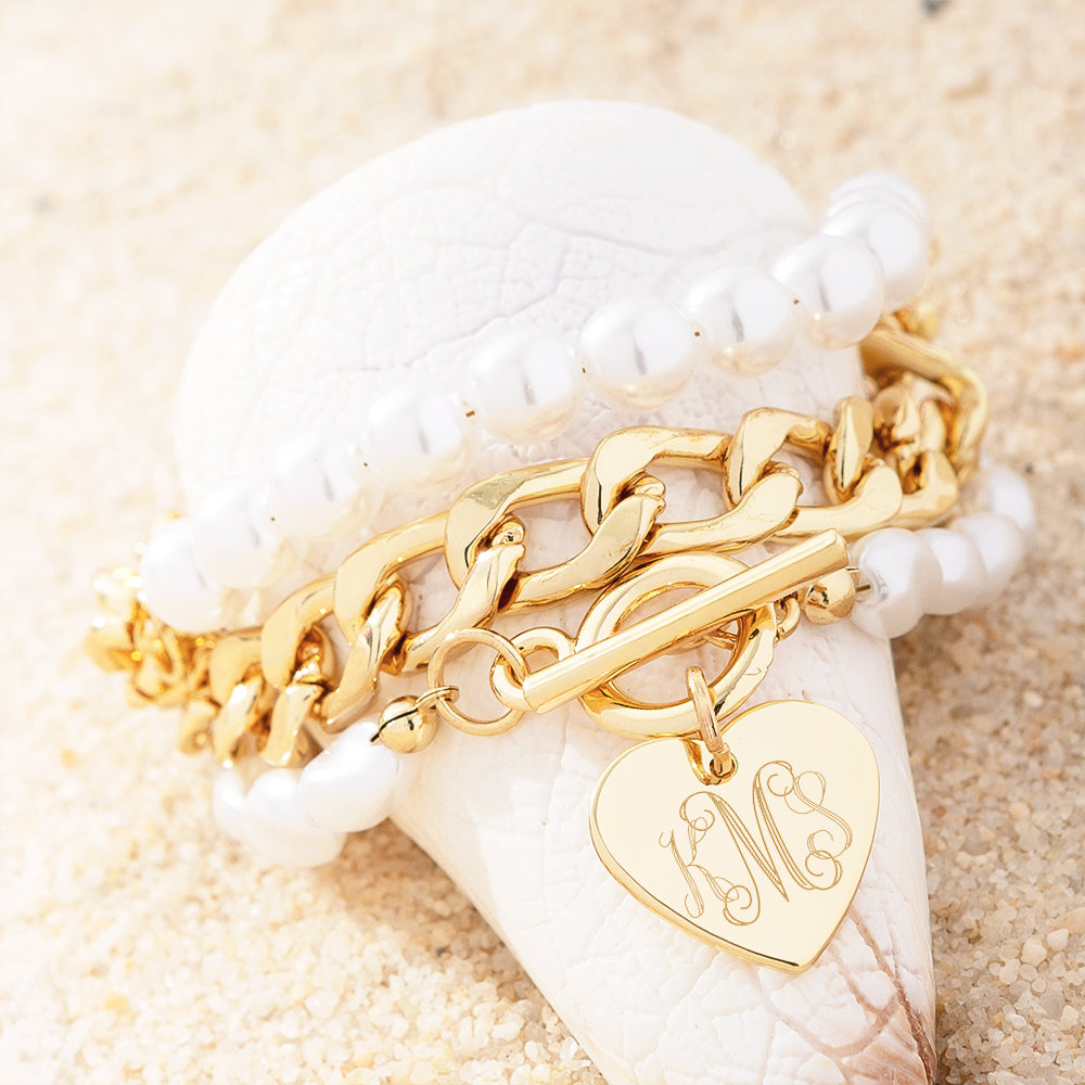Gold bracelet with a heart-shaped monogram charm on a seashell with sand background