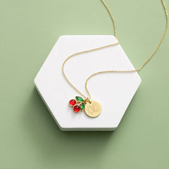 Gold necklace with red and green cherry charm on a white hexagonal stand against a light green background