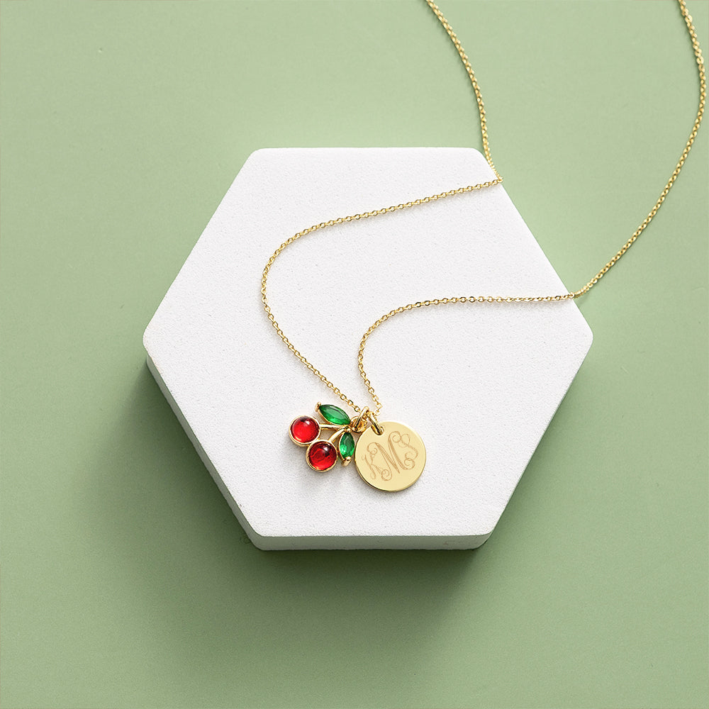 Gold necklace with red and green cherry charm on a white hexagonal stand against a light green background