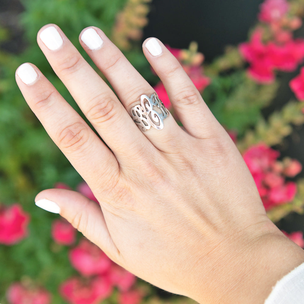 A sterling silver ring with a monogram cutout design worn on a hand with white nail polish, set against a blurred background of green foliage and pink flowers.