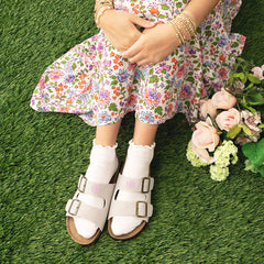 Woman wearing a floral dress and tan sandals on grass with flowers.