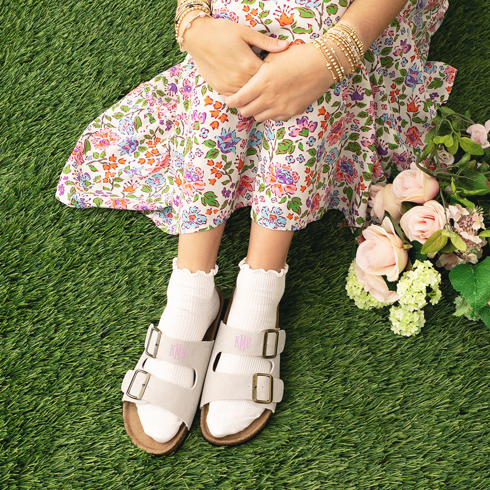 Woman wearing a floral dress and tan sandals on grass with flowers.
