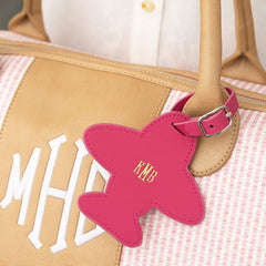 Pink airplane-shaped luggage tag with 'KMB' initials attached to pink and white seersucker weekender bag.