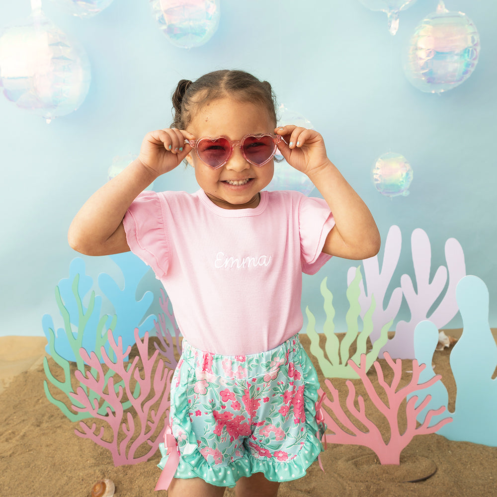 Child wearing pink sunglasses and a pink shirt with 'emma' on it, standing in front of underwater-themed decor.