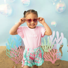 Child wearing pink sunglasses and a pink shirt with 'emma' on it, standing in front of underwater-themed decor.