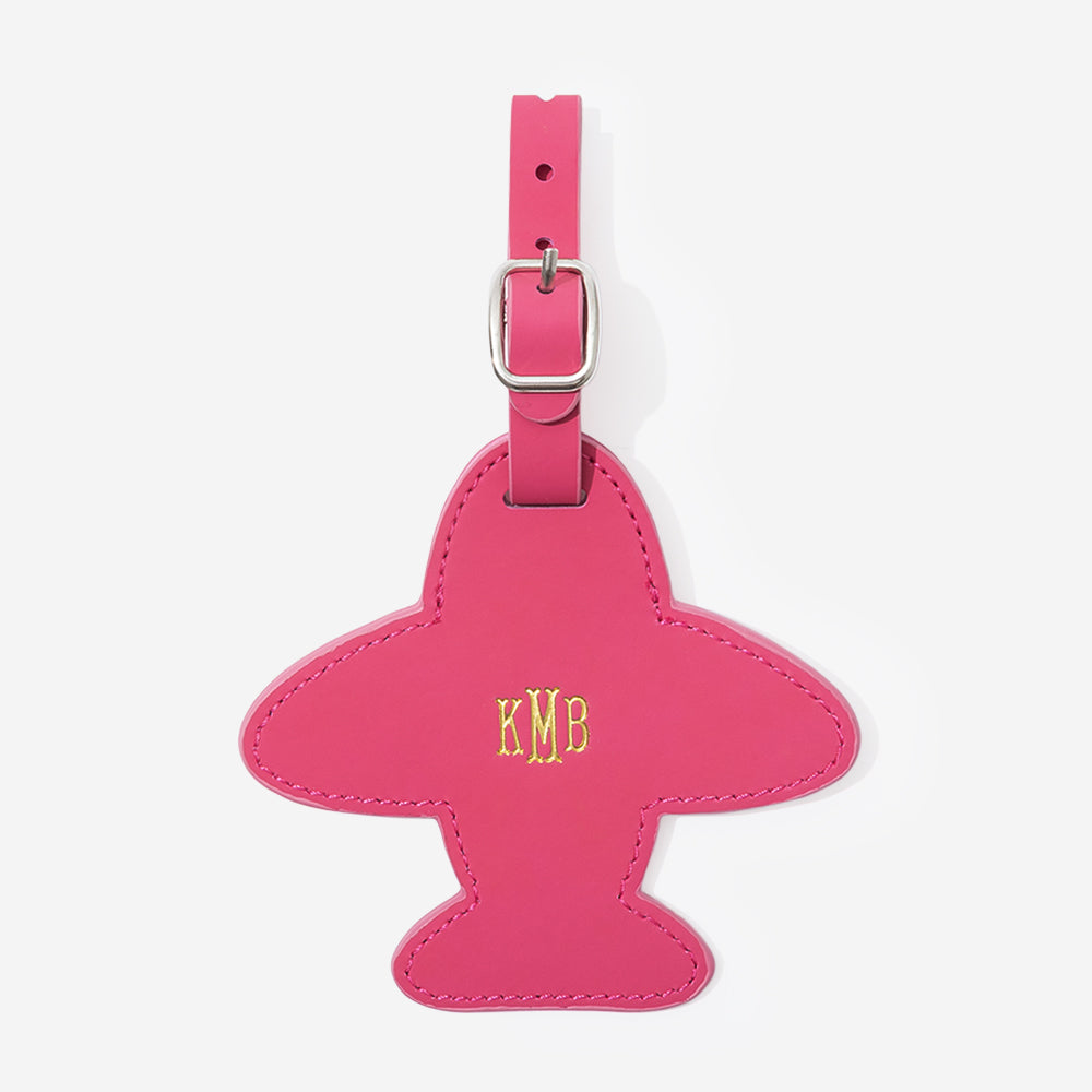Pink airplane-shaped leather luggage tag with gold monogram on a white background