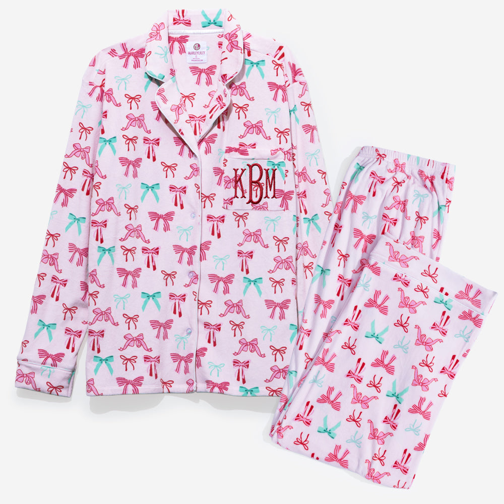 A two-piece pajama set featuring a long-sleeve button-up shirt and matching pants in light pink, both printed with a repeating pattern of pink and mint green bows. The shirt has a collar with dark red piping and a monogram 'KBM' embroidered on the left chest.