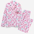 A two-piece pajama set featuring a long-sleeve button-up shirt and matching pants in light pink, both printed with a repeating pattern of pink and mint green bows. The shirt has a collar with dark red piping and a monogram 'KBM' embroidered on the left chest.