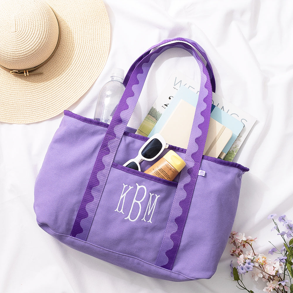 Purple tote bag with monogram 'KBM' containing a hat, bottle, sunglasses, and books on a white background.