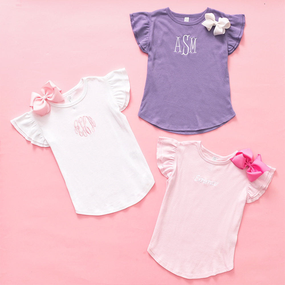 Three children's shirts with ruffled sleeves and bows on a pink background